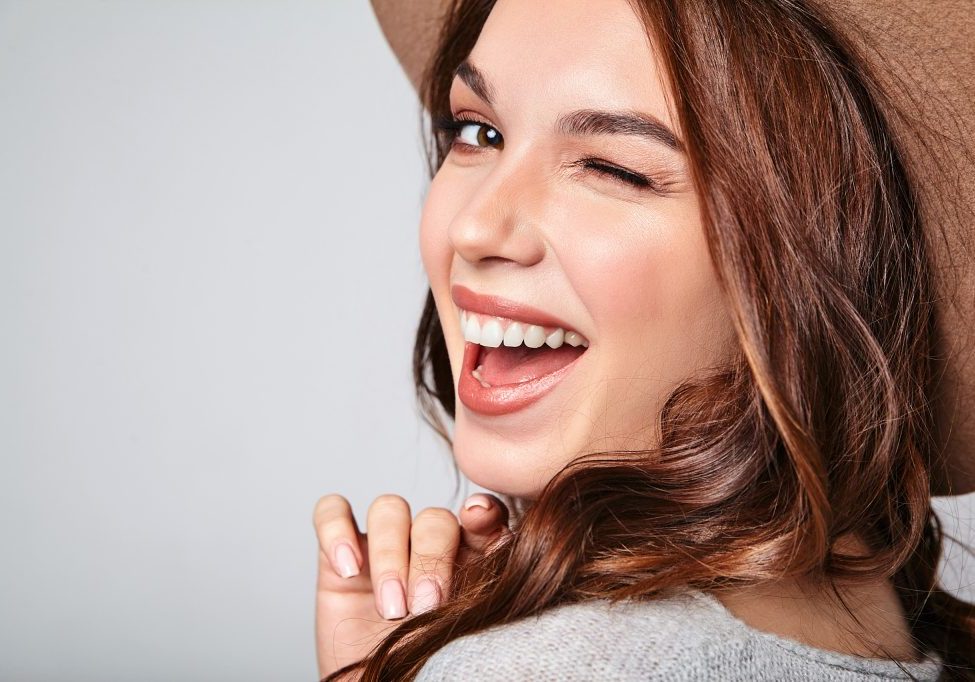 Portrait of young stylish laughing girl model in gray casual summer clothes in brown hat with natural makeup isolated on gray background. Looking at camera and winking